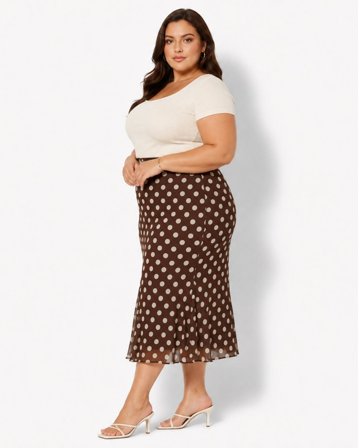 A woman modeling a brown midi skirt with a white polka dot pattern, paired with a cream off-the-shoulder top and white heeled sandals, against a plain white studio background.