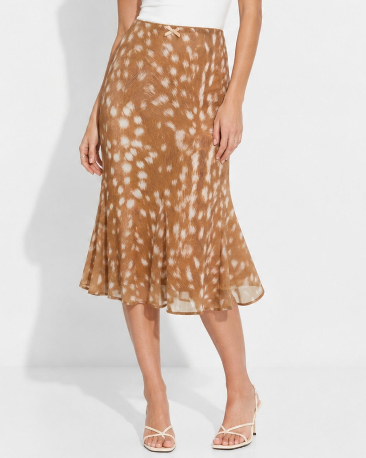 A woman models a brown midi skirt with a white tie-dye or watercolor splatter pattern, paired with a white top and white strappy heels against a white studio background.