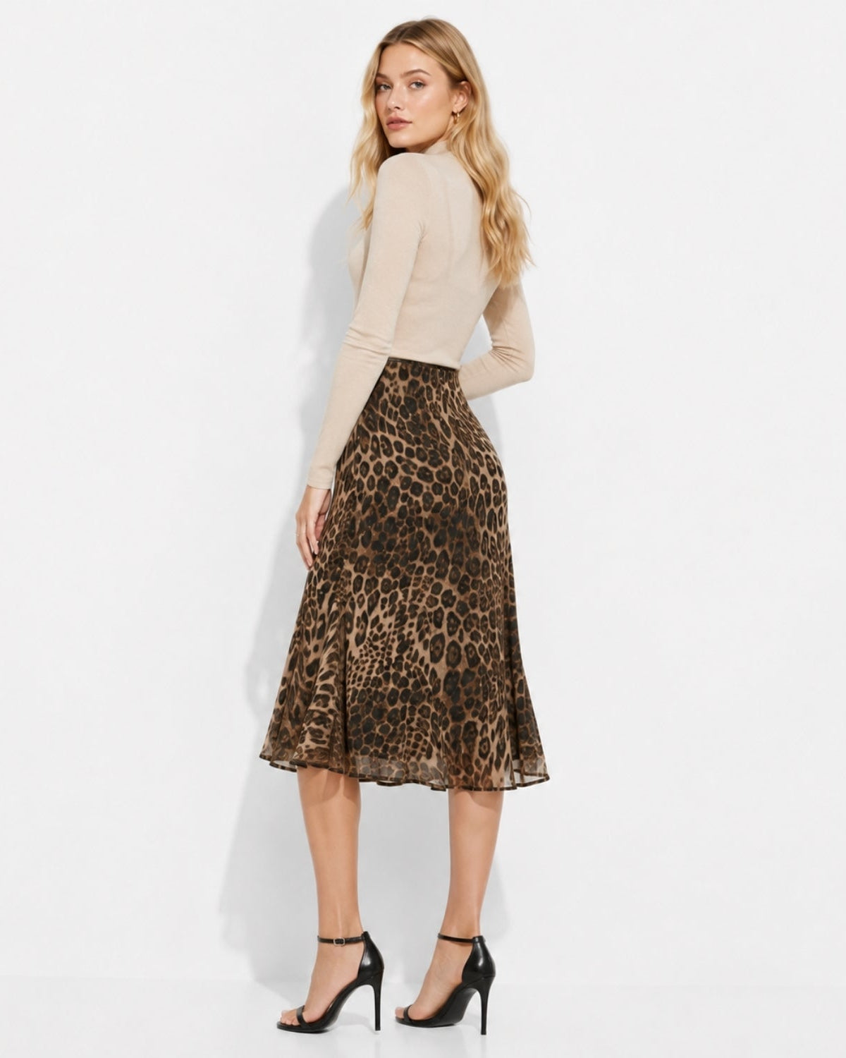 A woman modeling a leopard-print midi skirt with a beige long-sleeve top and black high-heeled sandals, photographed against a clean white studio background.