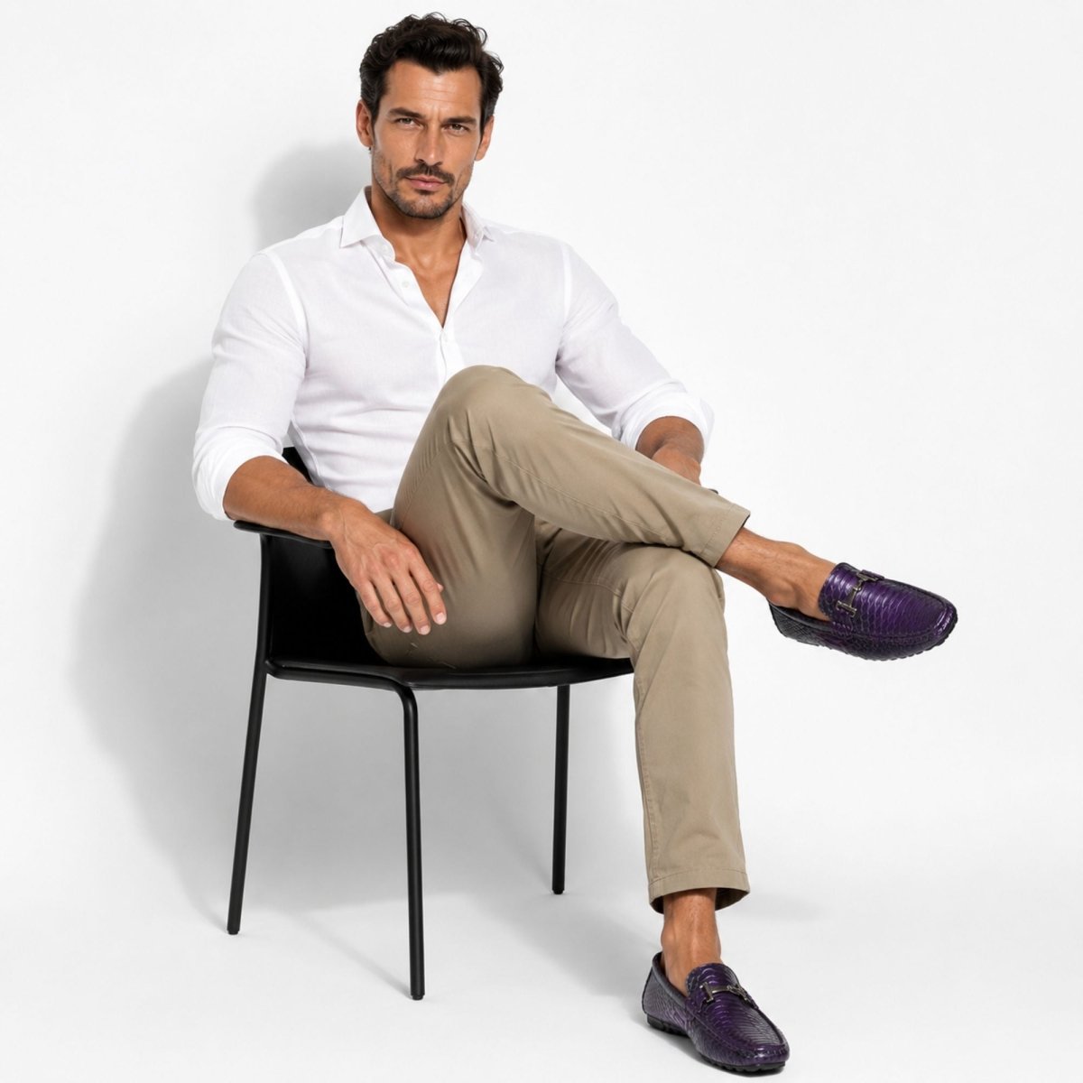A man wearing a white button-down shirt, beige trousers, and purple croc-embossed loafers sits relaxed in a black chair against a clean white studio background.