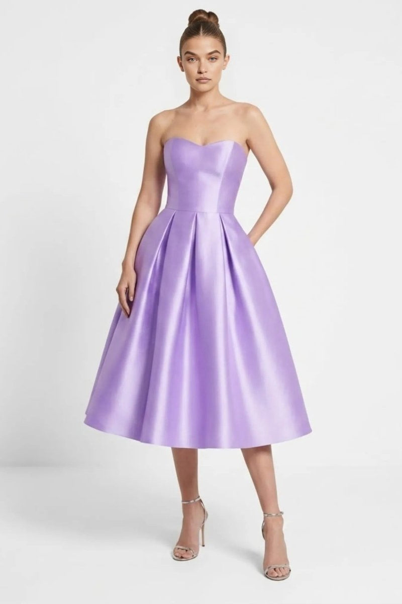 Front view of the Fenlo Form purple satin strapless pleated midi dress with pockets worn by a model in silver heels.