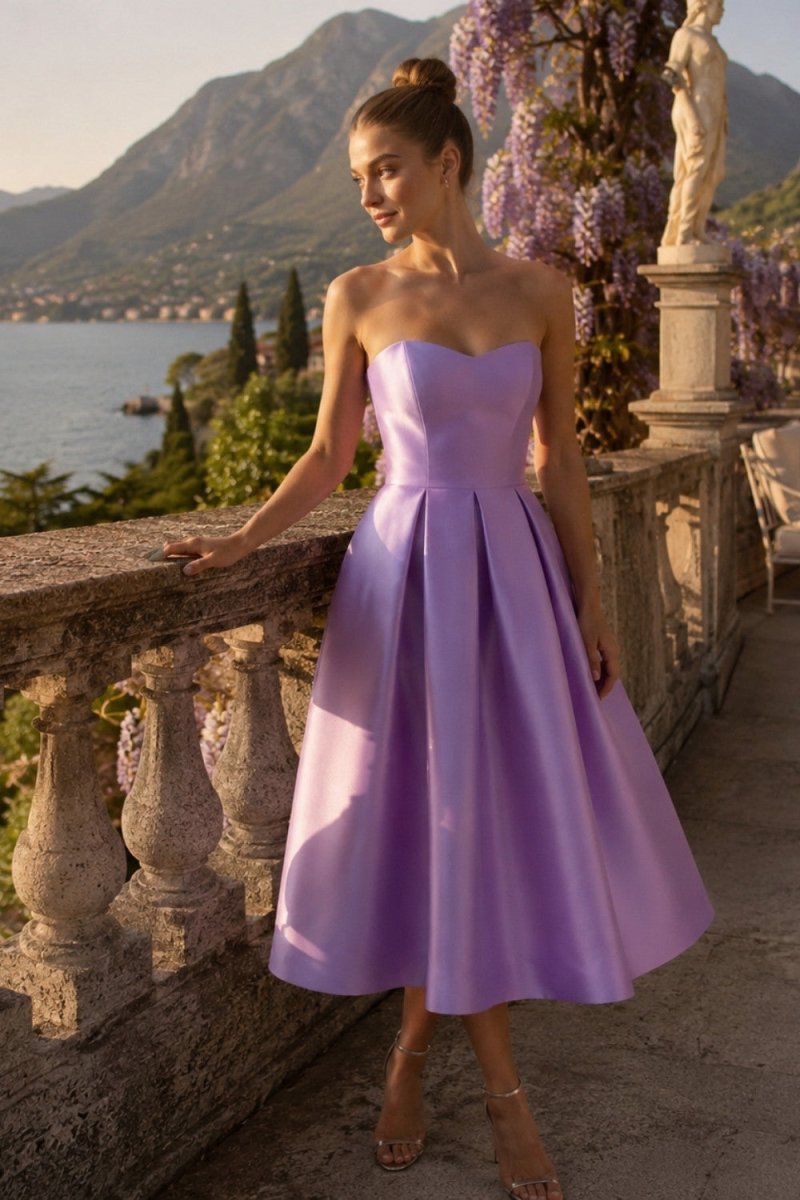 A woman wearing a strapless lavender midi dress with a sweetheart neckline and a full skirt, posing on an outdoor terrace with a lake and mountains in the background.