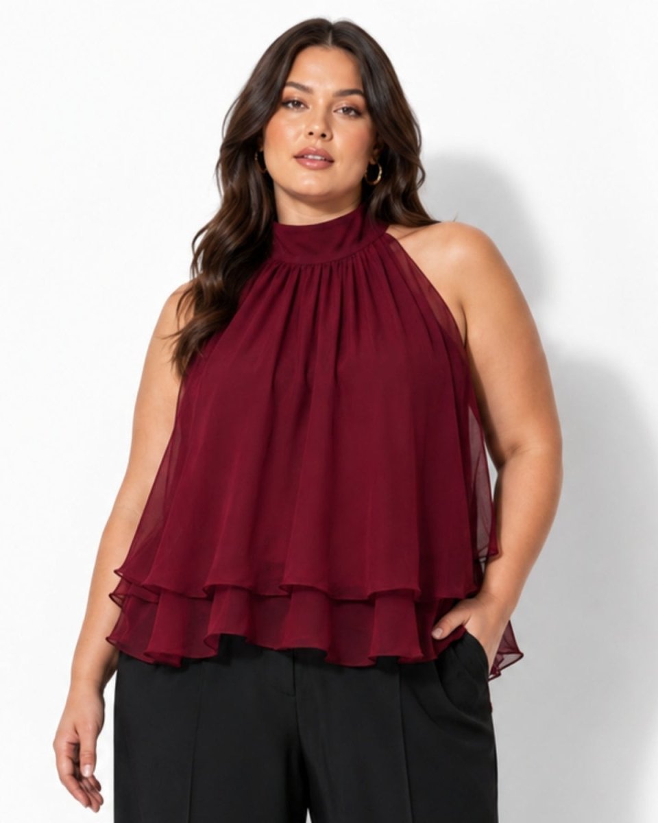 A woman modeling a sleeveless high-neck blouse in deep burgundy with a gathered, pleated ruffled hem, paired with black pants, against a plain white studio background.