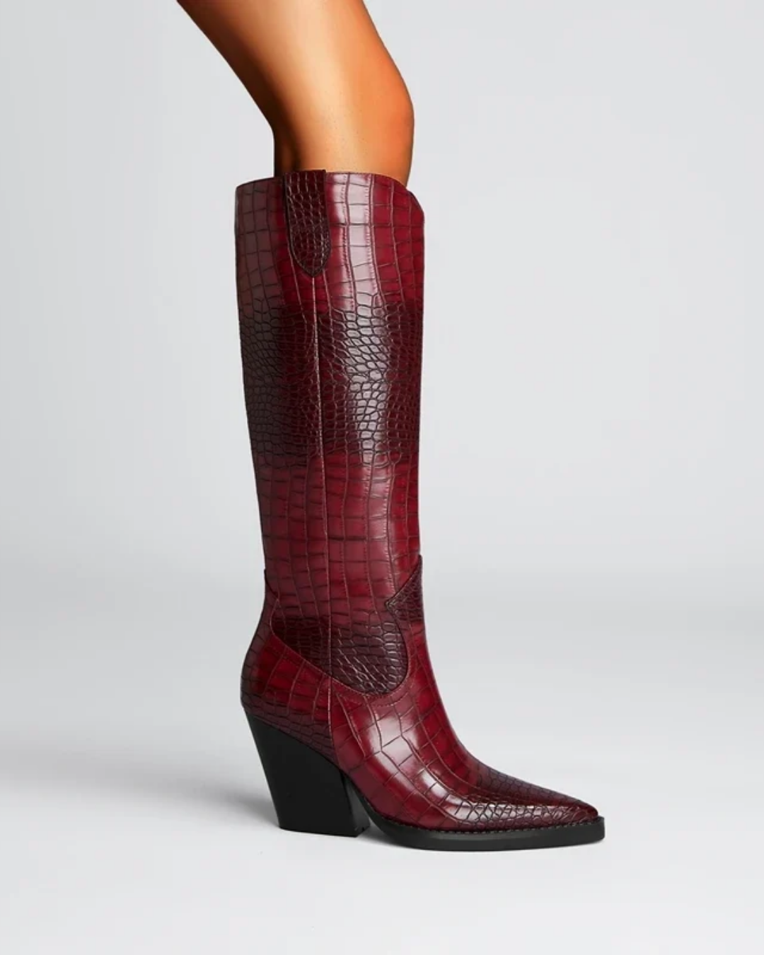 A single knee-high boot with a dark red gradient and crocodile-embossed texture, shown on a model's leg against a light gray studio background.