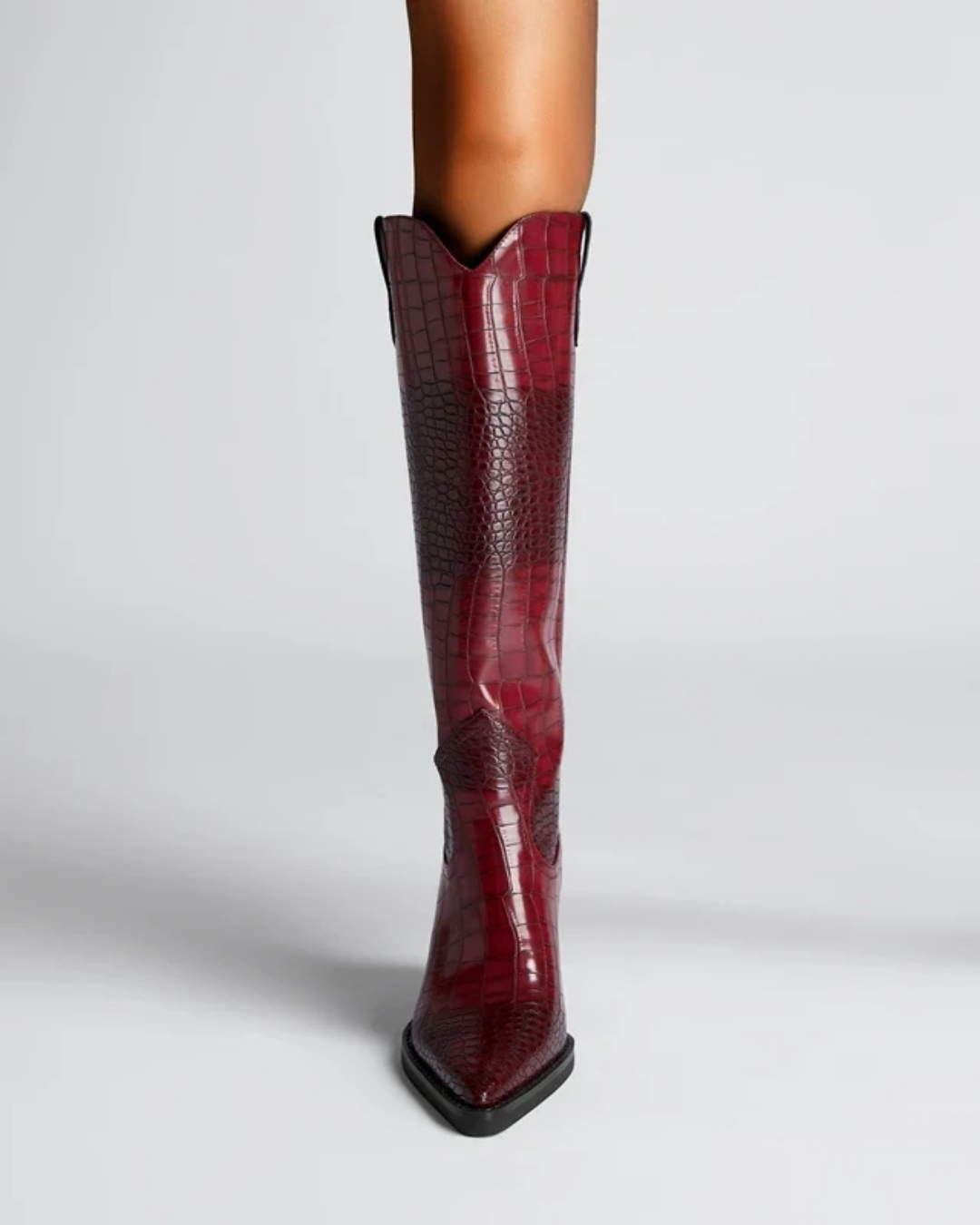 A single knee-high boot in glossy embossed crocodile-pattern leather, shown on a model's leg against a light gray studio background.
