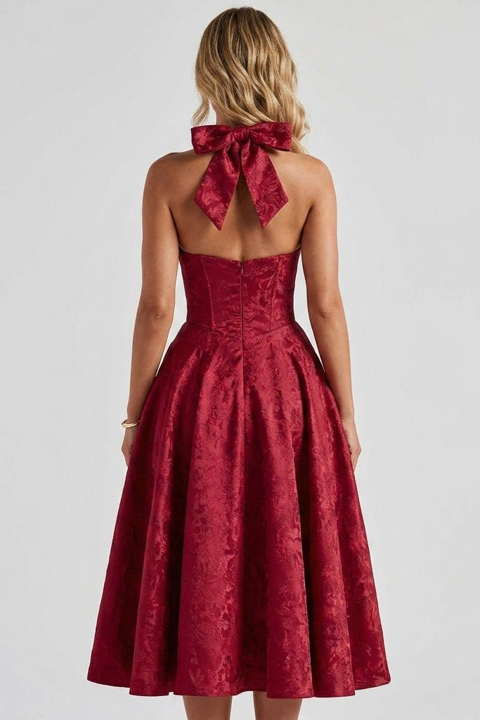 Back view of a woman wearing the Kouvr red jacquard formal dress highlighting the open back design, large dramatic neck bow, and flared A-line skirt.