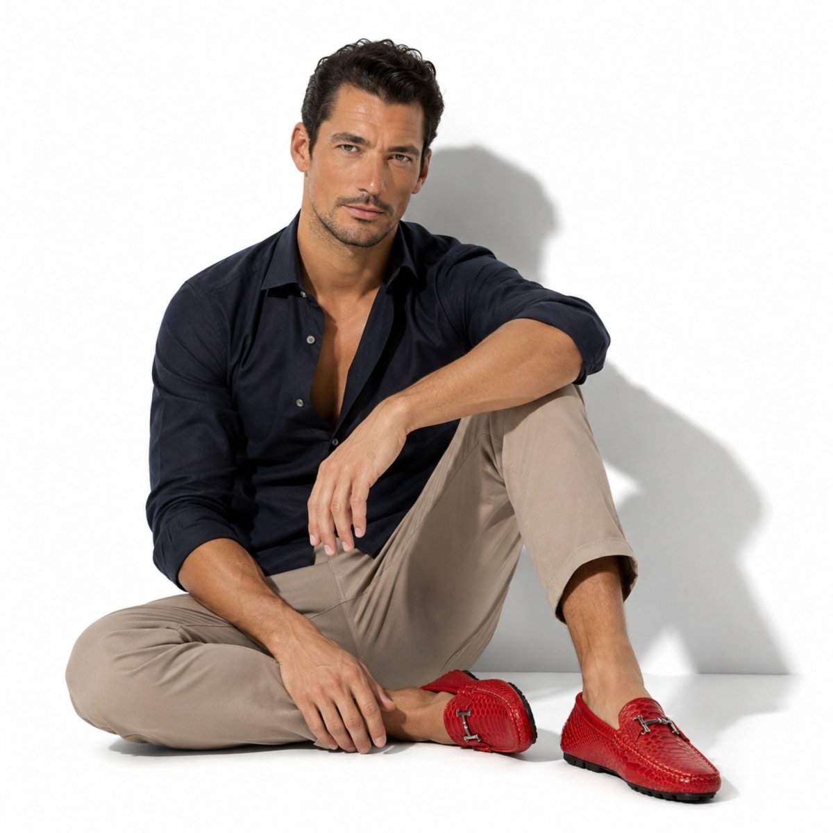 A man sits cross-legged on a white studio floor wearing a dark navy button-down shirt, beige trousers, and bright red loafers. A strong shadow is cast on the white background.