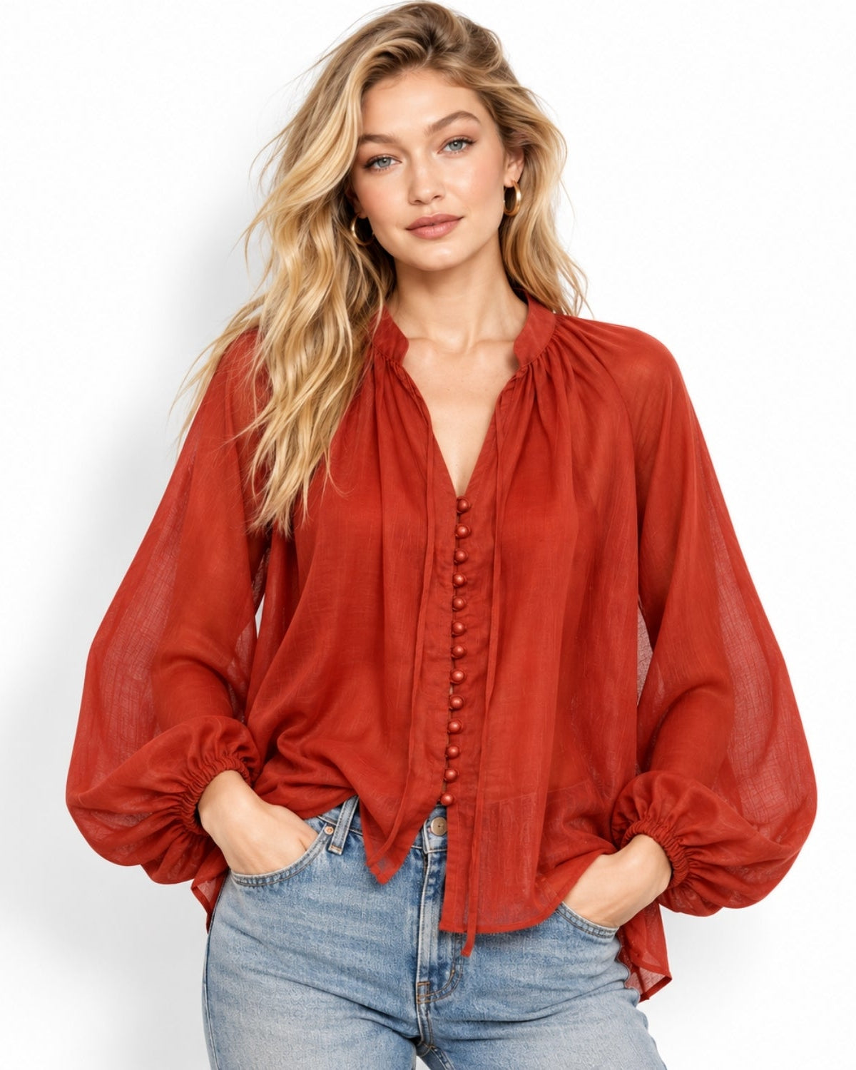 A woman modeling a rust-orange sheer long-sleeve blouse with a gathered V-neck and button front, paired with light-wash blue jeans against a plain white studio background.