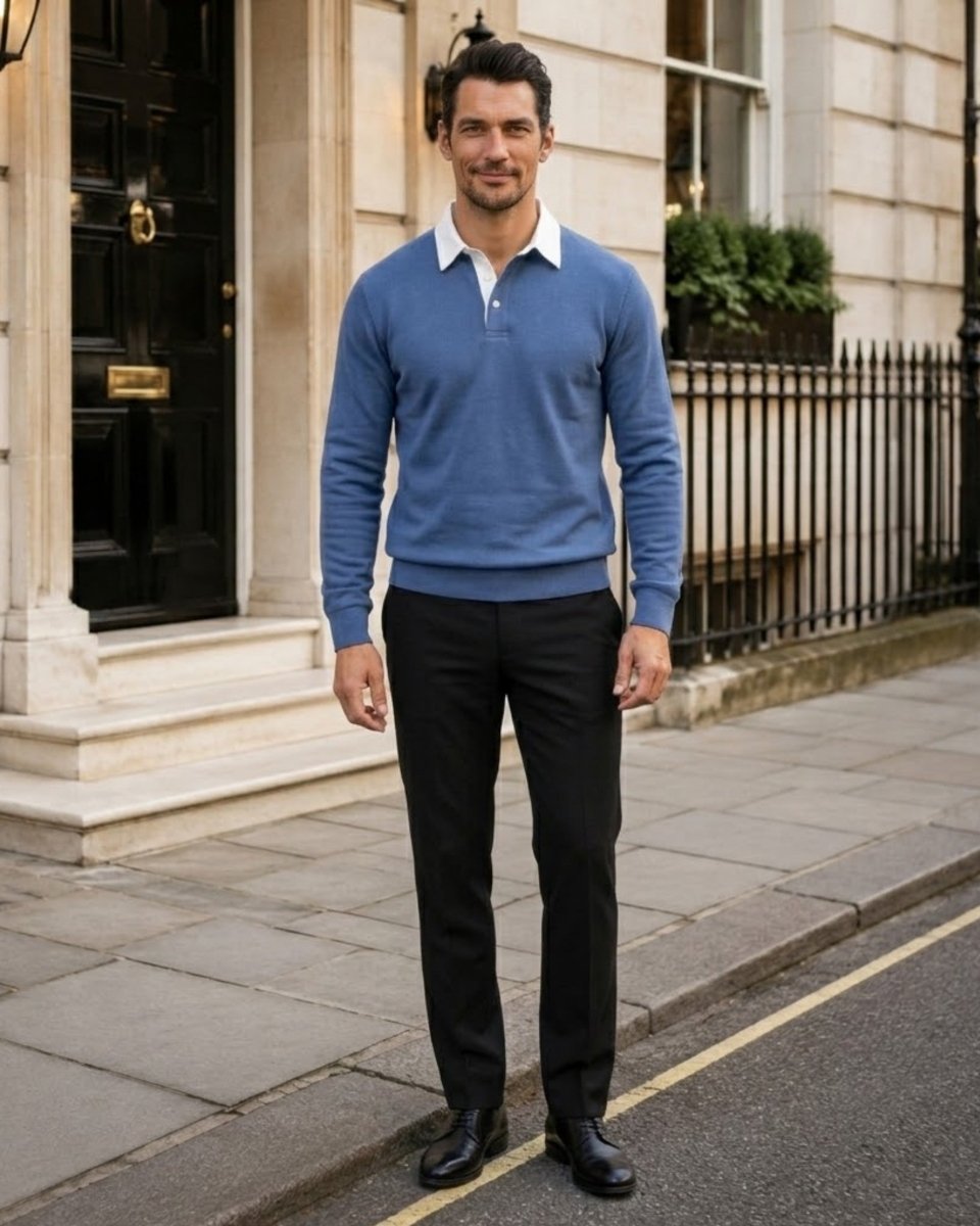 A man standing on a city sidewalk wearing a blue long-sleeve polo-style sweater over a white collared shirt, black trousers, and black shoes, with a building facade in the background.