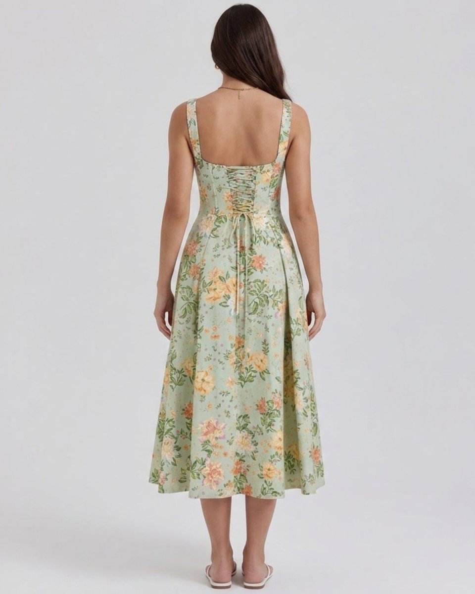 A woman viewed from the back wearing a sleeveless midi dress with a light sage green floral print, tied waist, and square neckline, set against a plain light gray studio background.