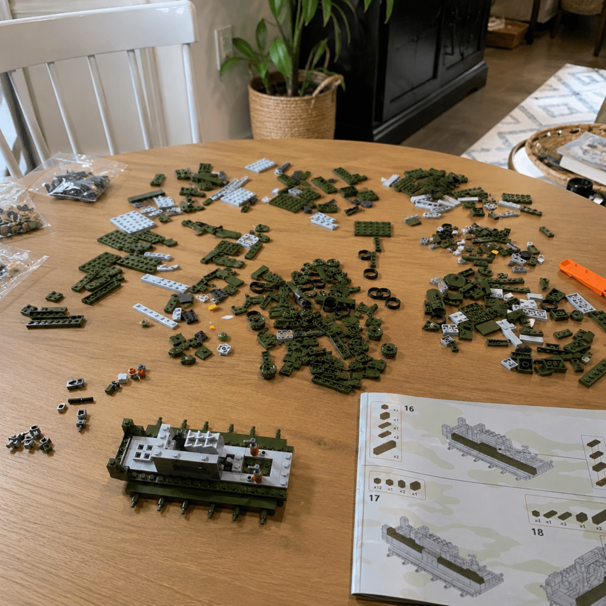 A wooden table covered with scattered building-brick pieces and an instruction sheet, with a partially assembled green and gray brick model in the foreground. A chair, potted plant, and indoor room are visible in the background.