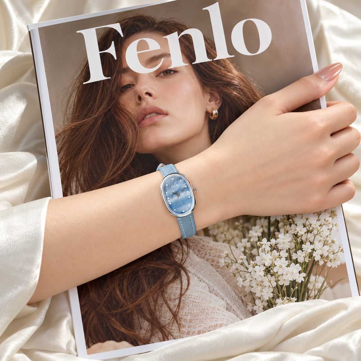 A woman's hand wearing a light blue watch with an oval face and blue strap, resting on a magazine cover featuring a woman with the brand name 'Fenlo' at the top, set against a soft cream satin fabric background.