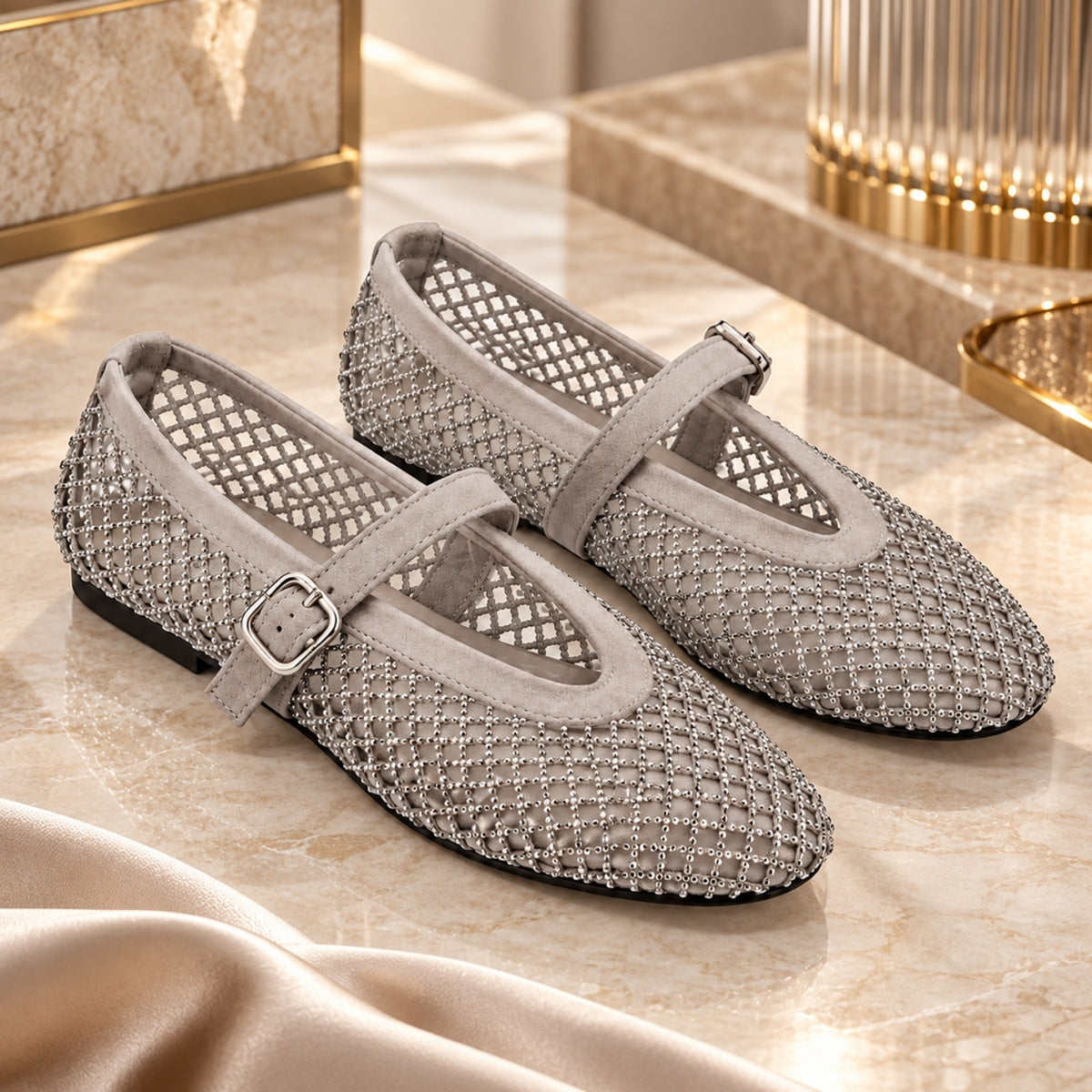 A pair of women's Mary Jane flats in a light gray suede and silver rhinestone mesh, displayed on a beige marble surface with elegant gold-toned decor in the background.