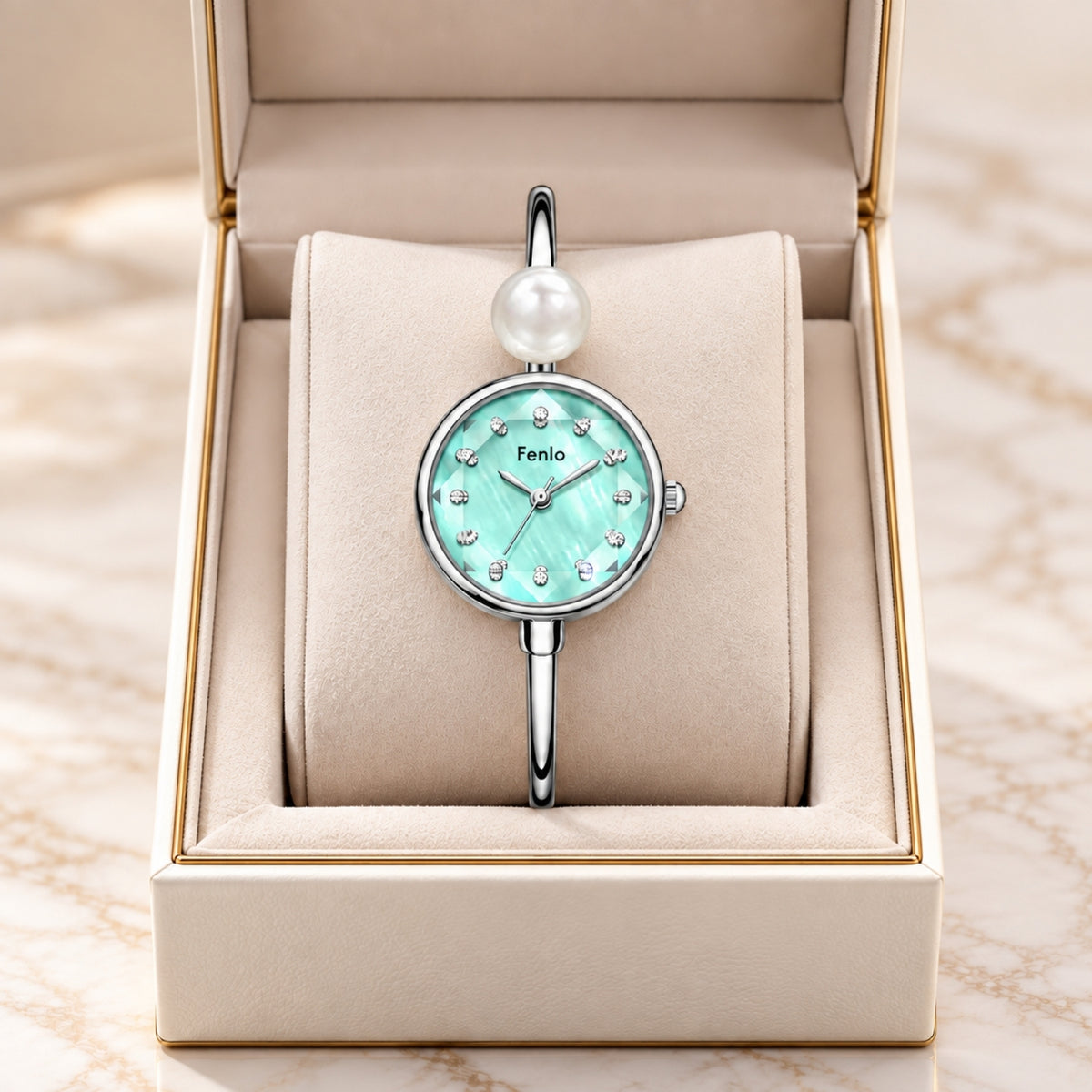 A silver-tone bangle watch with a round mint green dial, crystal hour markers, and a single white pearl at the top, displayed inside an open beige jewelry box on a marble surface.