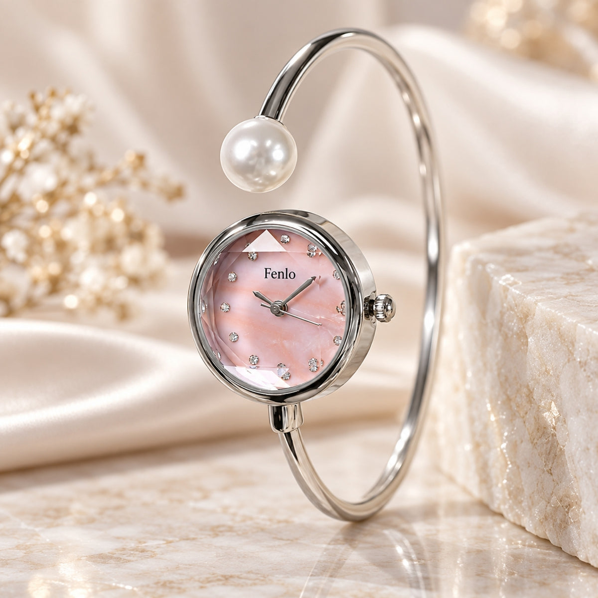 A silver-toned bangle watch with a round pink dial, rhinestone hour markers, a pearl-like bead at the top, and the brand name 'Fenlo' on the dial, styled on a marble surface with soft cream fabric and decorative flowers.