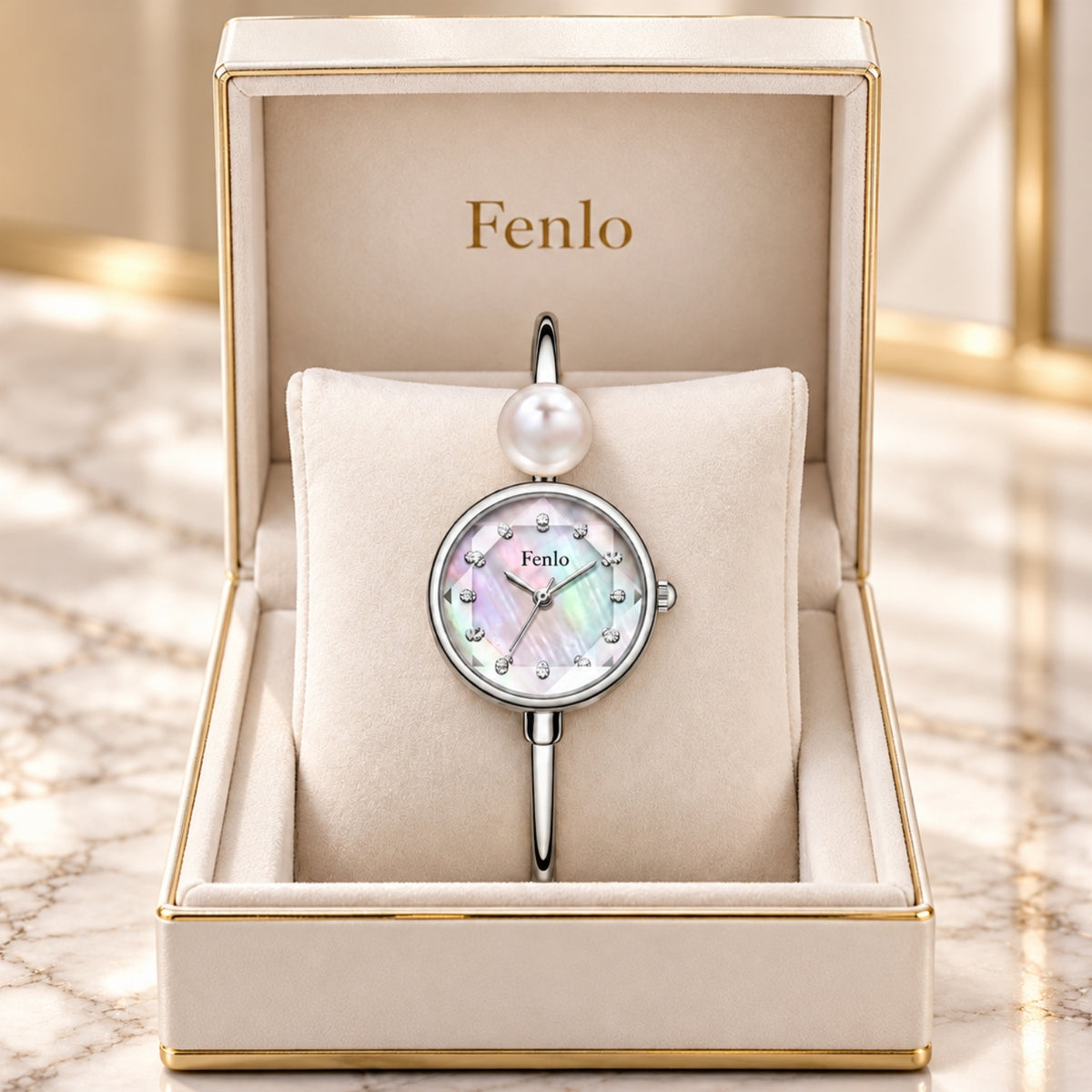 A Fenlo wristwatch with a round mother-of-pearl dial and a single pearl at the top of the strap, presented in an open cream jewelry box with gold trim on a marble surface.