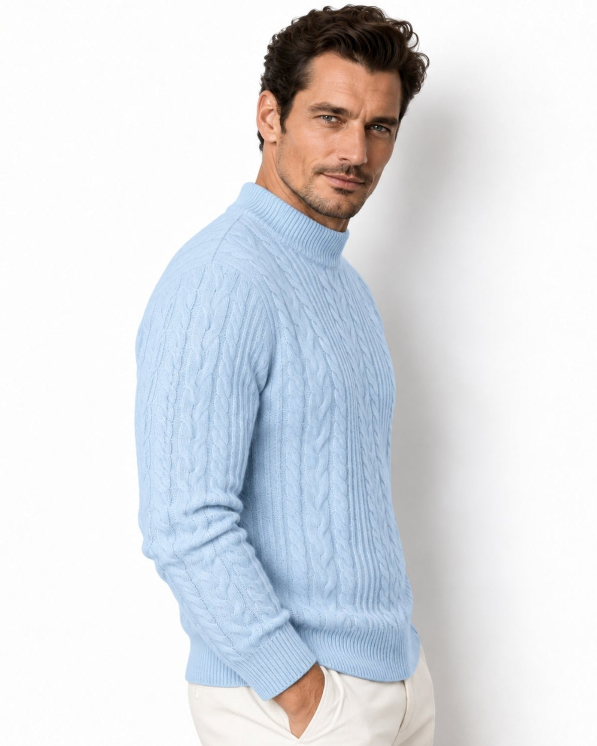 A man wearing a sky blue cable-knit mock neck sweater and off-white trousers, posing against a clean white studio background.