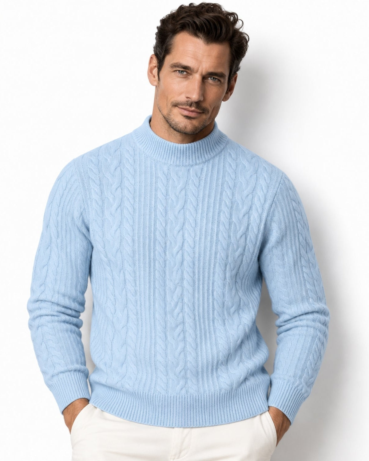 A man wearing a light blue cable-knit mock neck sweater and off-white trousers, standing against a plain white studio background.