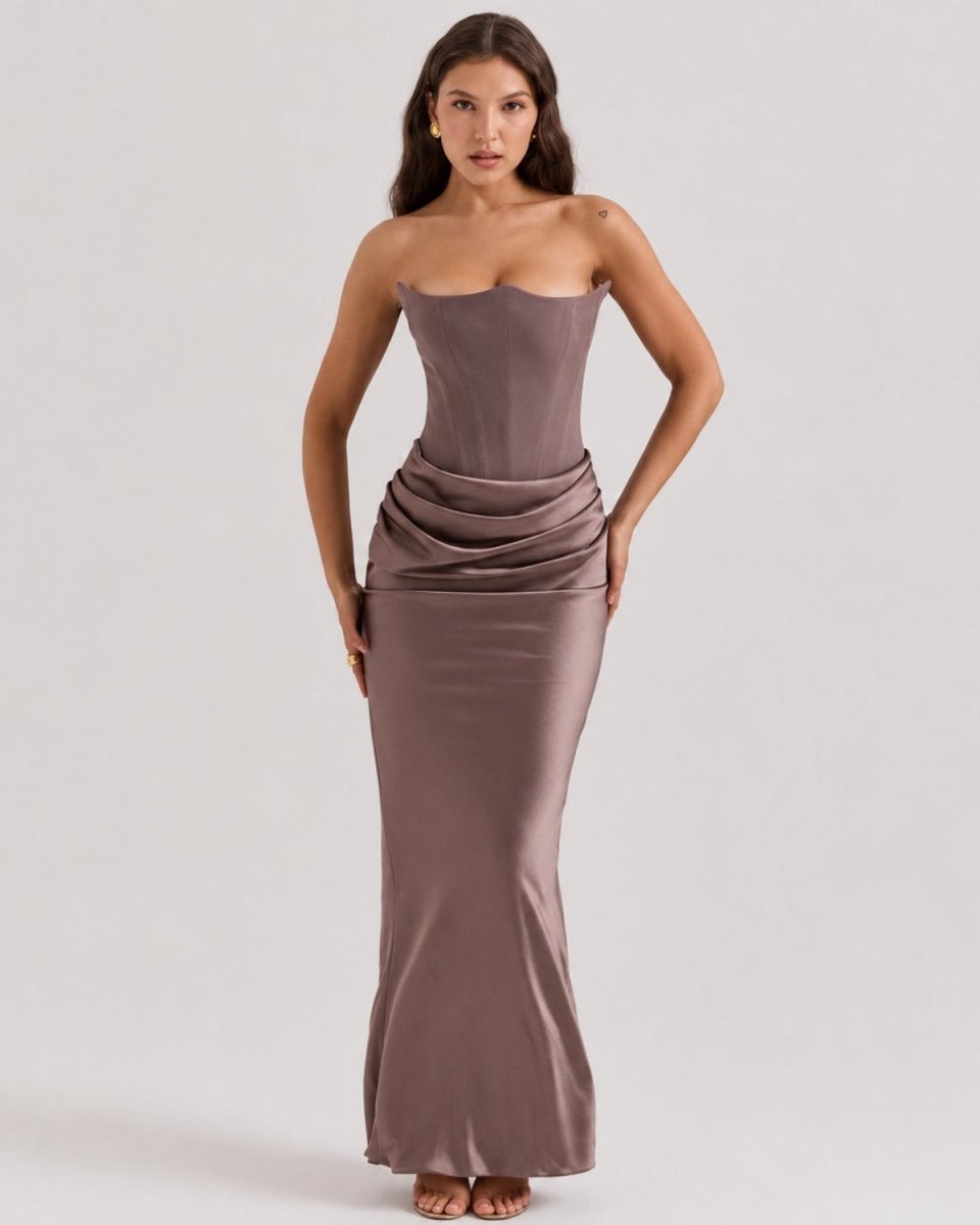 A woman modeling a strapless, floor-length satin gown in a muted taupe-brown shade against a plain light gray studio background.