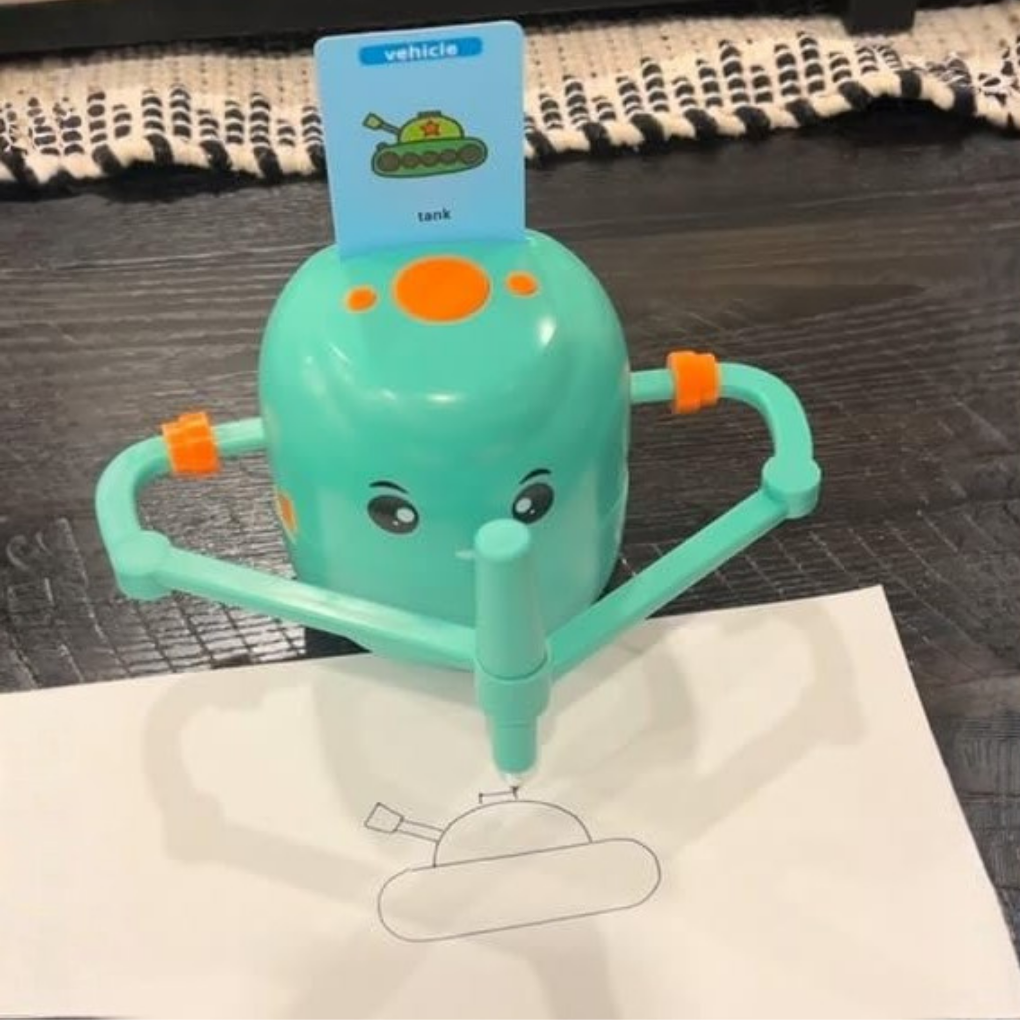 A teal drawing toy for kids with a cartoon face, featuring arm-like supports and a card labeled 'vehicle' and 'tank' propped behind it, drawing on a white sheet of paper over a dark wood surface.