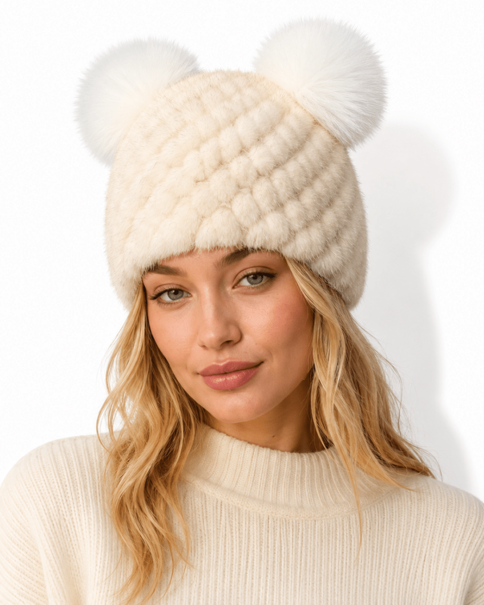 A woman wearing a cream-colored faux fur beanie with two large fluffy pom-poms on top, paired with a matching cream ribbed sweater against a plain white background.