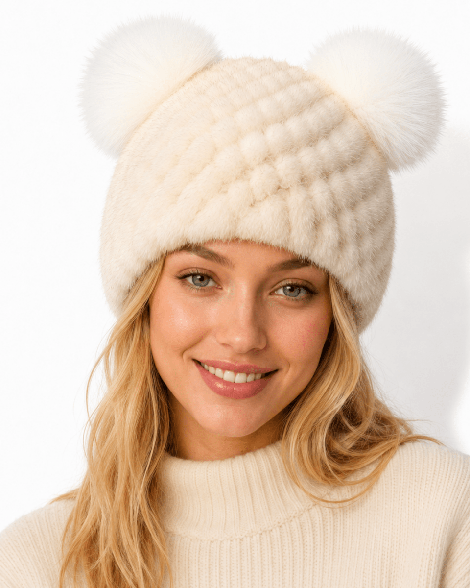 A woman wearing a cream-colored knit beanie with two fluffy pom-poms on top, paired with a matching cream ribbed sweater, against a clean white background.