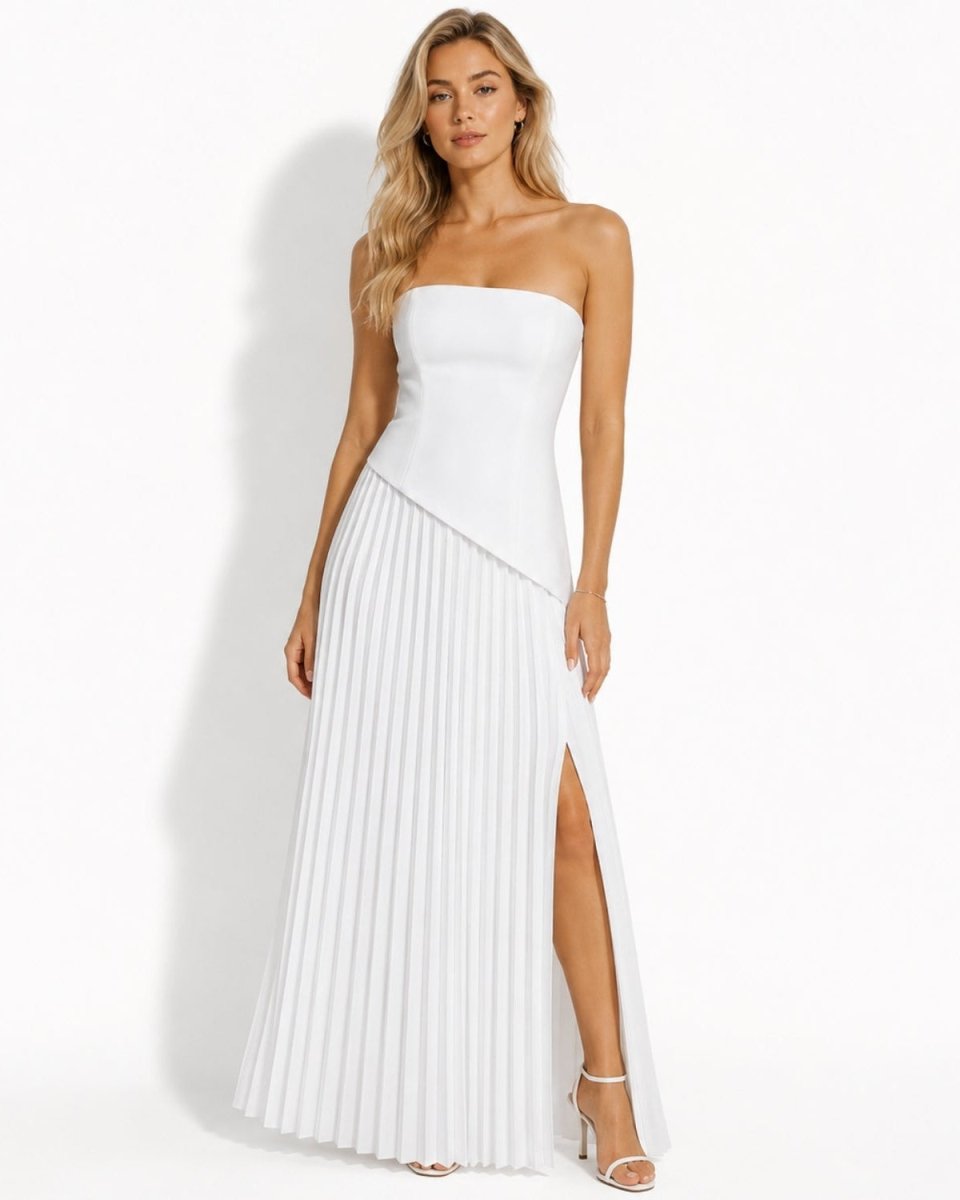 A woman modeling a strapless white maxi dress with a structured corset-style bodice, asymmetrical draped waist detail, and a pleated skirt with a front slit, photographed against a clean white studio background.