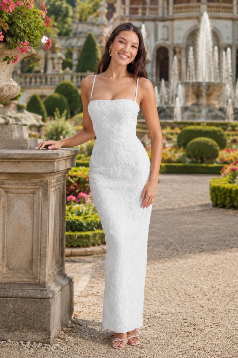 A woman wearing a fitted white lace spaghetti-strap maxi dress stands outdoors in a formal garden setting with a stone pedestal in the foreground and a fountain in the background.