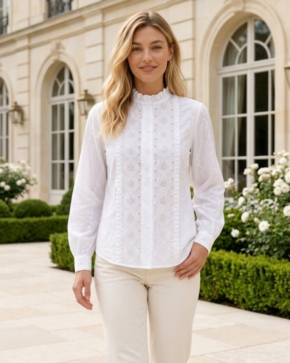 A woman wearing a white long-sleeve eyelet blouse with a high ruffled collar and cream trousers, standing outdoors in front of a grand beige building with arched windows and green hedges.