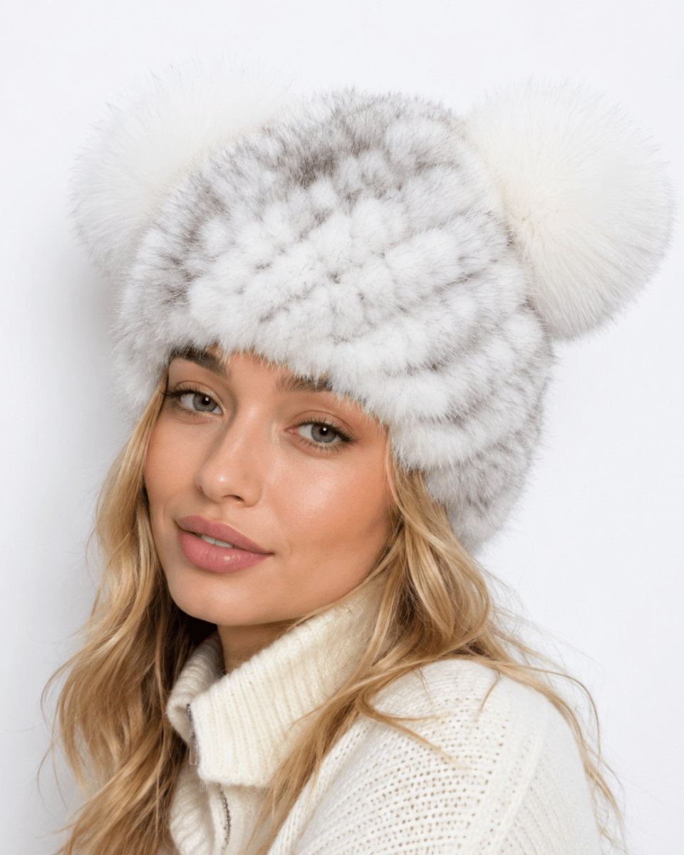 A woman wearing a fluffy white and gray striped fur hat with two large cream faux-fur pom-poms, paired with a cream knit sweater against a plain white background.