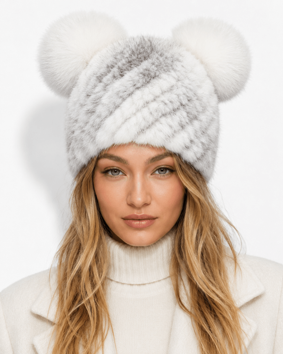 A woman wearing a white faux fur beanie with two large pom-poms on top, paired with a cream turtleneck sweater, against a plain white background.
