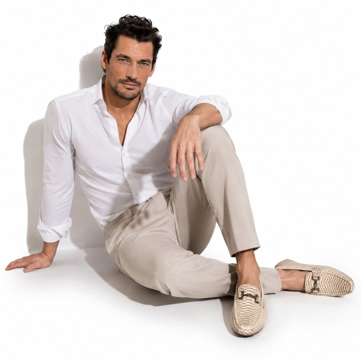 A man sitting on a white studio floor wearing a white button-down shirt, beige trousers, and beige woven loafers with dark metal hardware.