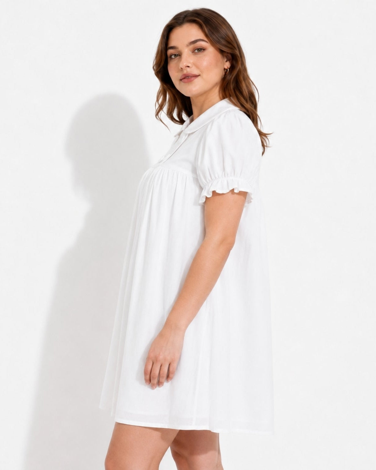 A woman modeling a white short-sleeve babydoll dress with a ruffled collar and sleeve cuffs, standing against a plain white studio background.