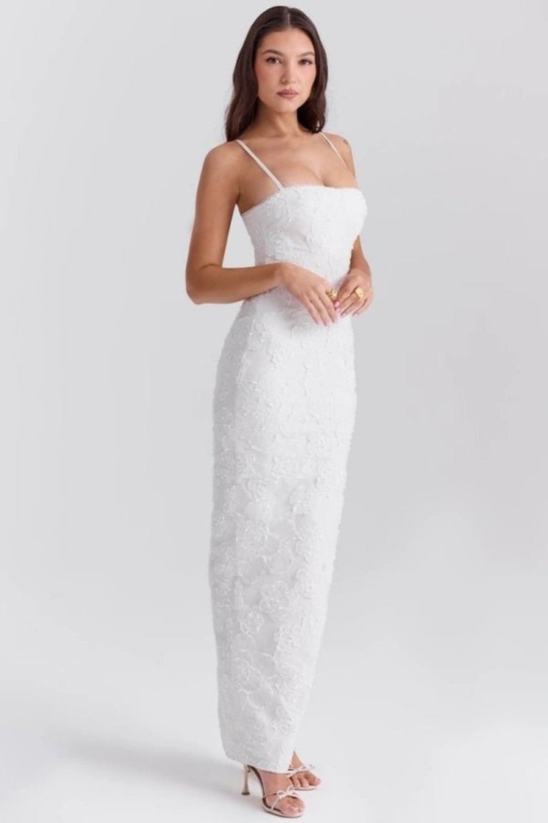 A woman modeling a white strapless lace maxi dress with spaghetti straps and a fitted silhouette, paired with nude high-heeled sandals, against a light gray studio background.