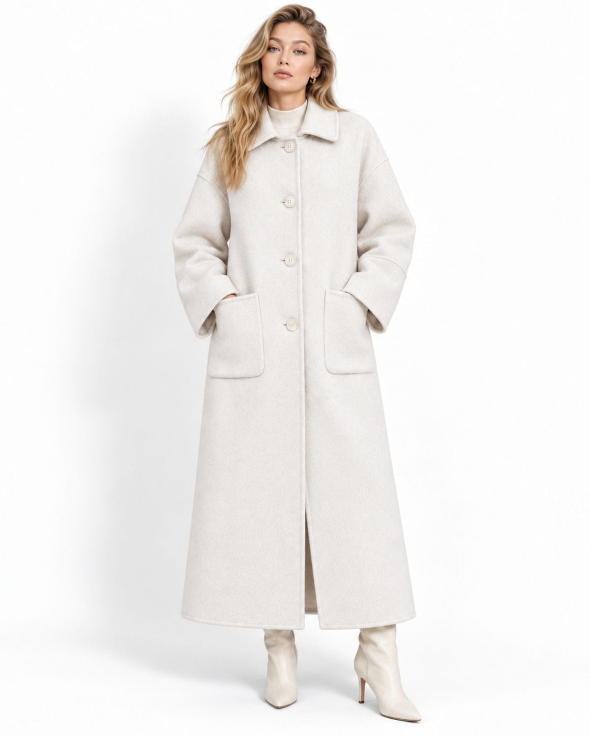 A woman modeling a long, oversized white-colored wool coat with front buttons and large patch pockets, paired with matching cream ankle boots, against a plain white studio background.