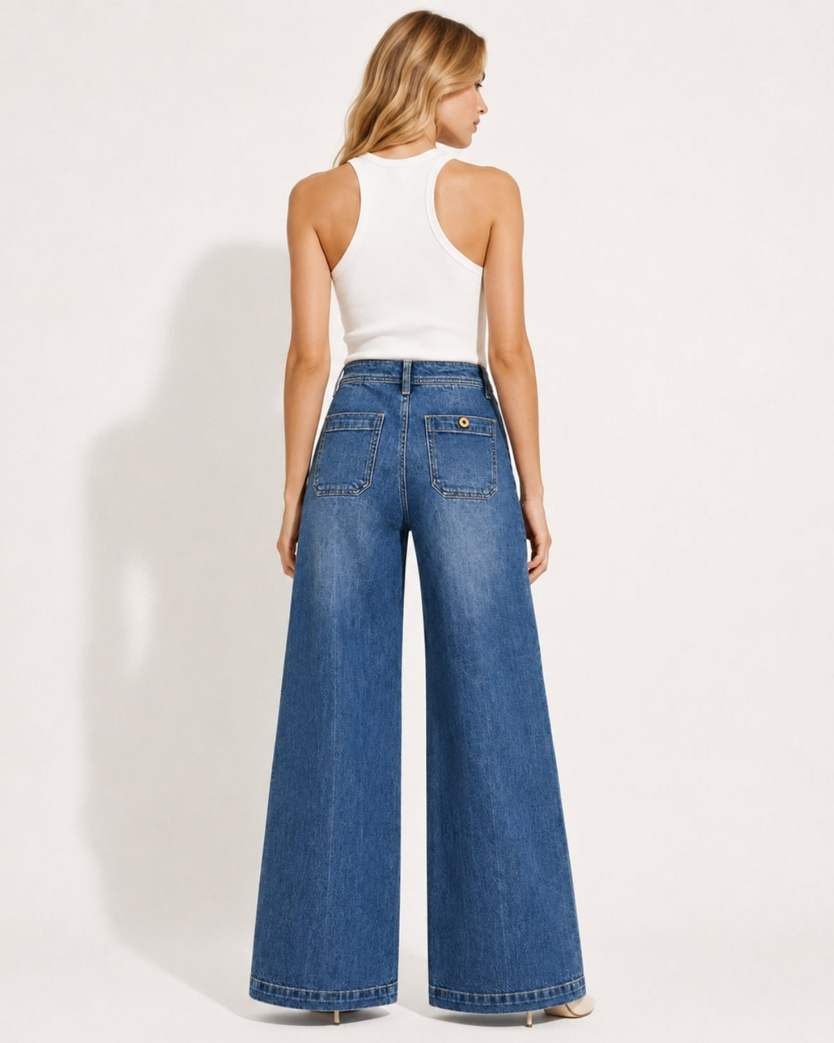 Back lifestyle view of model wearing high-waisted jeans highlighting the tailored seat, back patch pockets with brass button detail, and wide leg hem.
