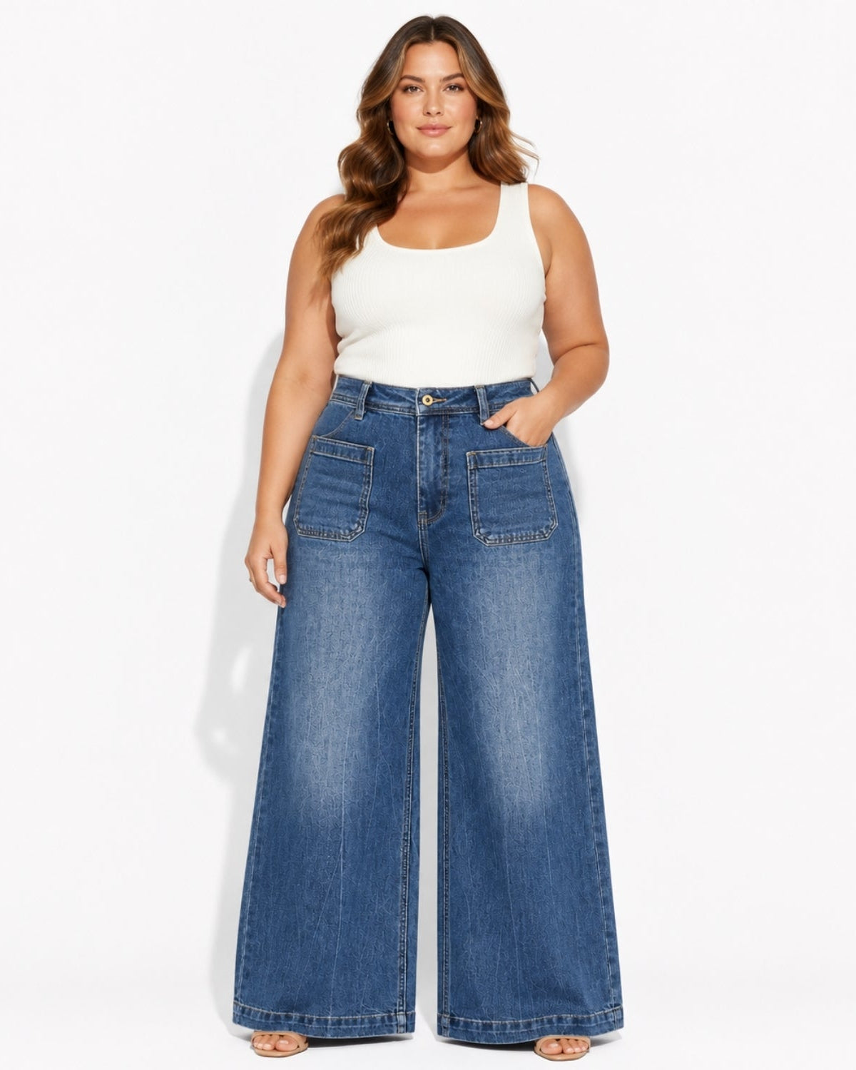 A woman modeling a pair of high-waisted, wide-leg blue denim jeans with a white tank top, standing against a plain white studio background.