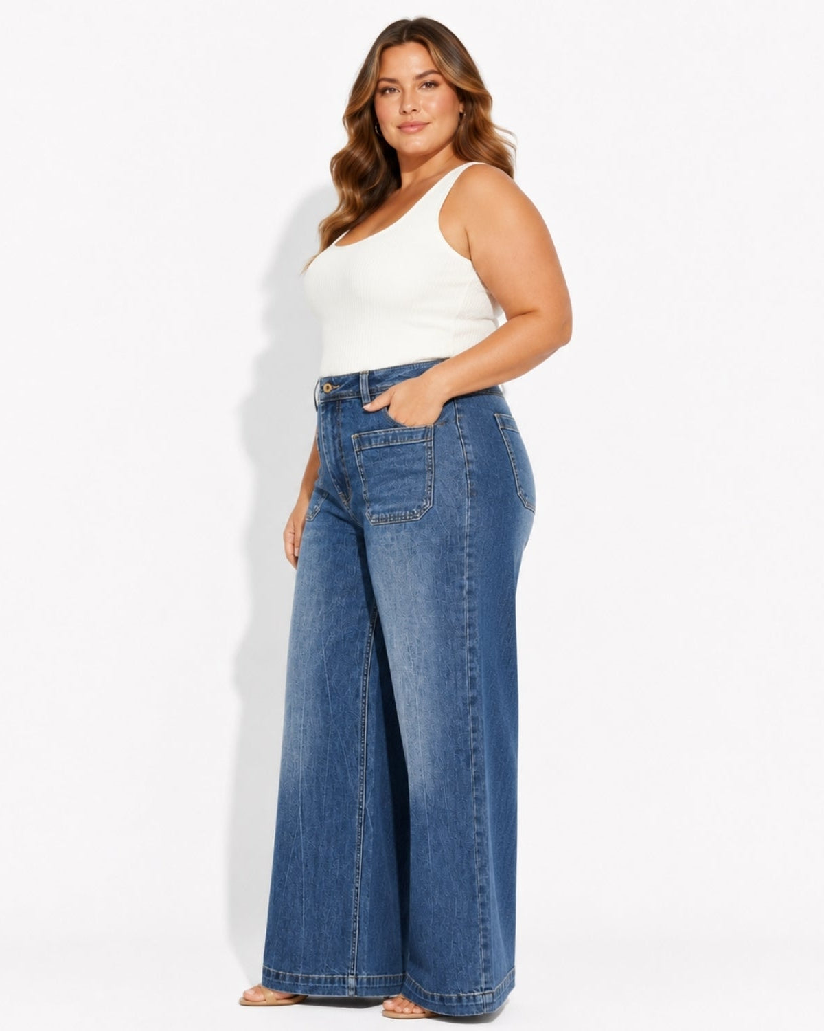 A woman modeling a pair of high-waisted, wide-leg blue denim jeans with a white ribbed tank top, standing against a plain white studio background.