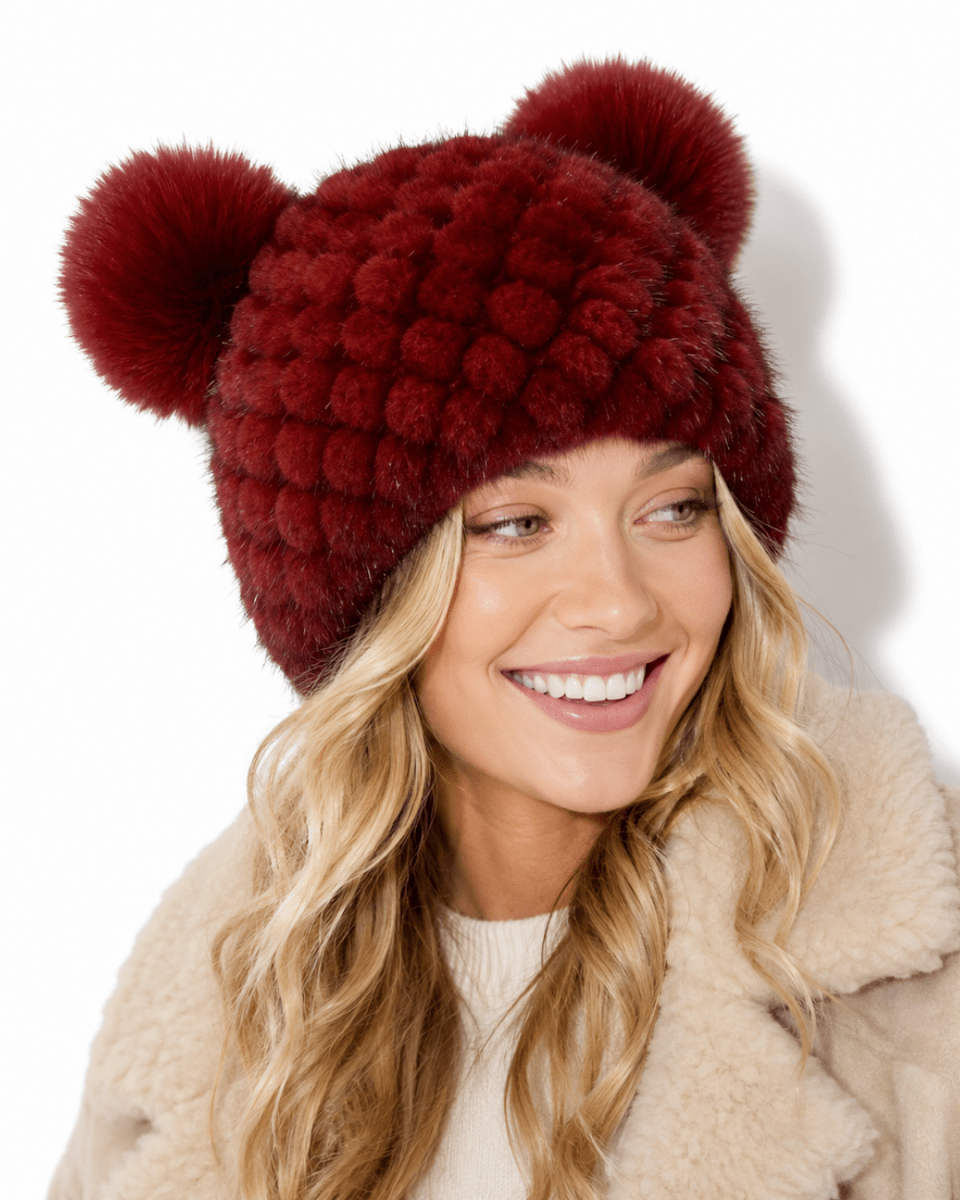 A smiling blonde woman wearing a burgundy faux fur beanie with two large pom-poms, paired with a cream shearling coat against a white background.