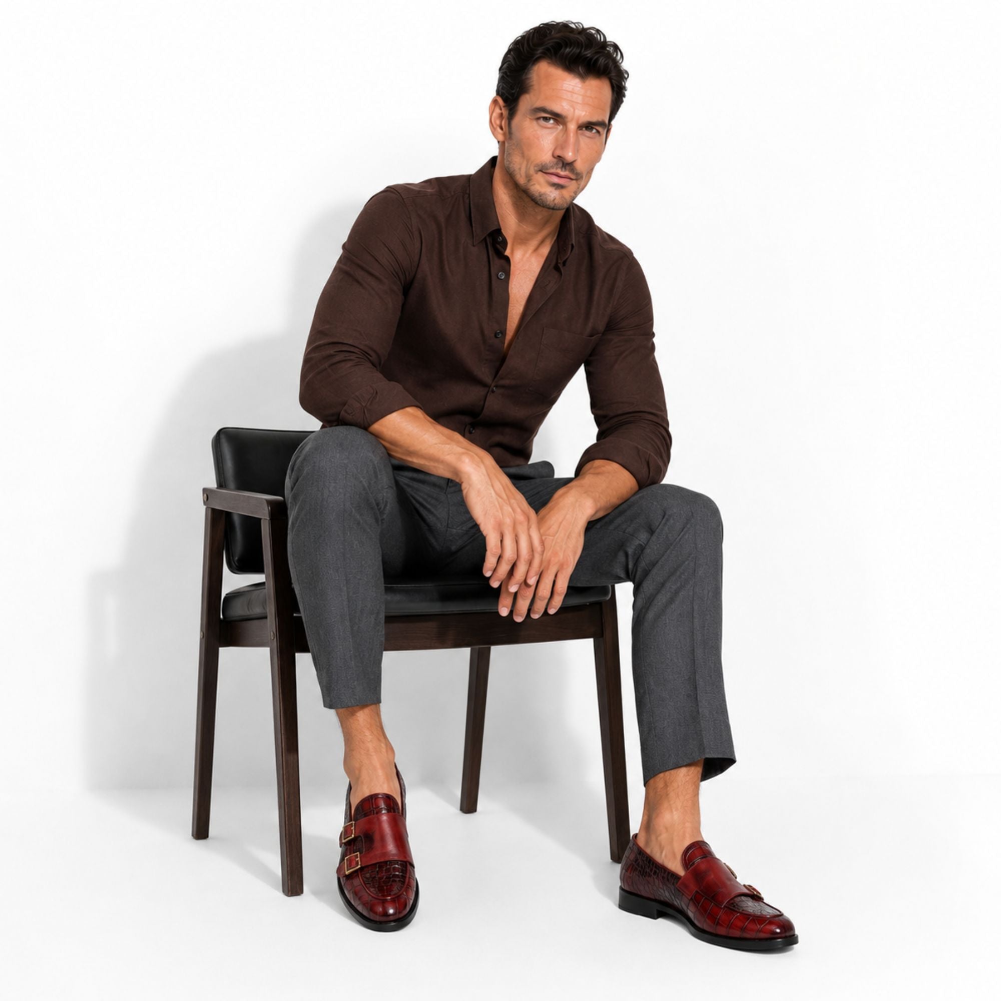 A man sits on a dark wooden chair wearing a brown button-down shirt, dark gray trousers, and dark red double-strap loafers against a plain white studio background.