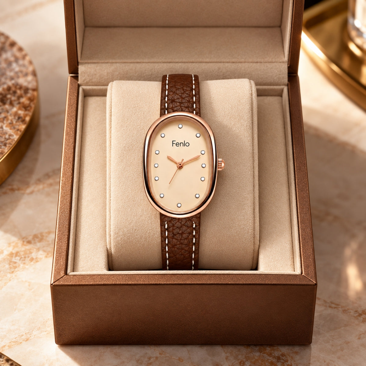 An oval Fenlo wristwatch with a rose gold case and a brown textured strap, presented inside an open brown gift box on a beige cushion. The watch face is cream with crystal hour markers.