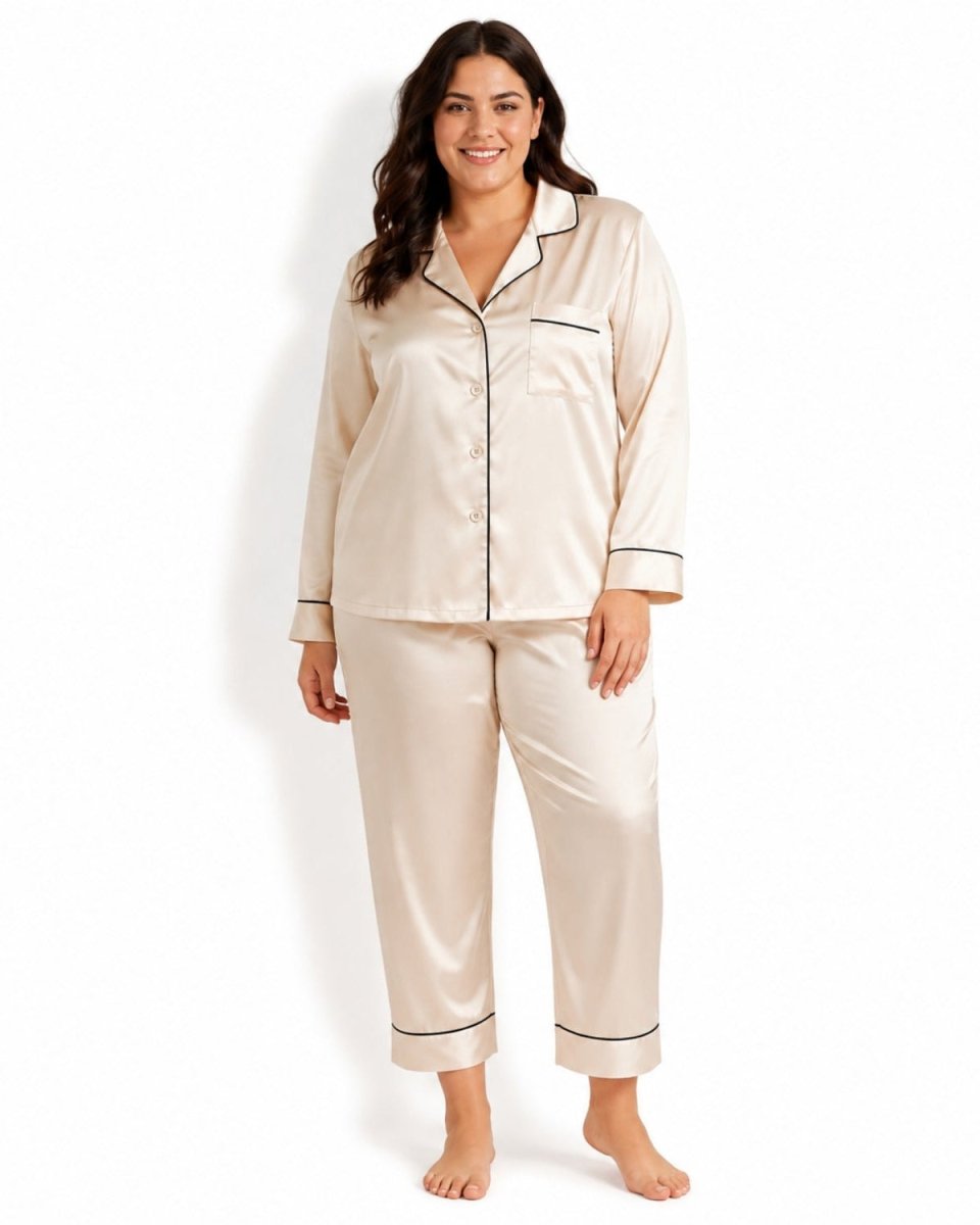 A woman modeling a two-piece satin pajama set in champagne beige with black piping, consisting of a long-sleeve button-front pajama top and matching pants, against a plain white background.