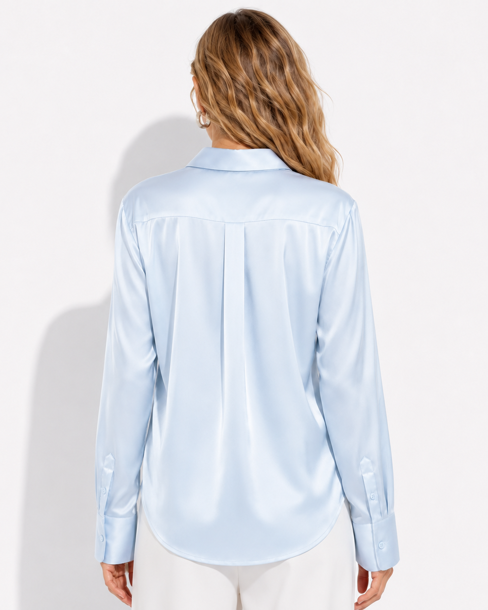 A woman viewed from the back wearing a light blue satin long-sleeve button-up shirt and white pants, set against a plain white studio background.