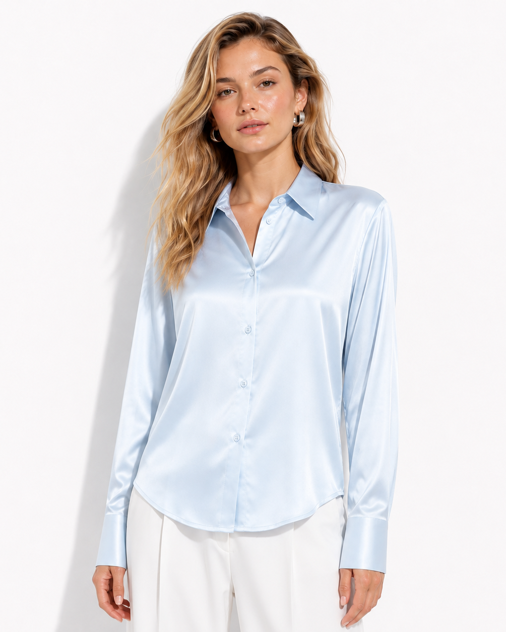 A woman modeling a light blue satin button-up blouse with white trousers, standing against a plain white studio background.
