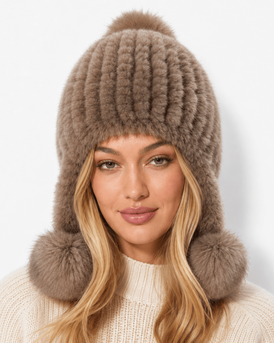 A woman wearing a ribbed brown faux fur beanie with ear flaps and pom-poms, photographed against a clean white background.
