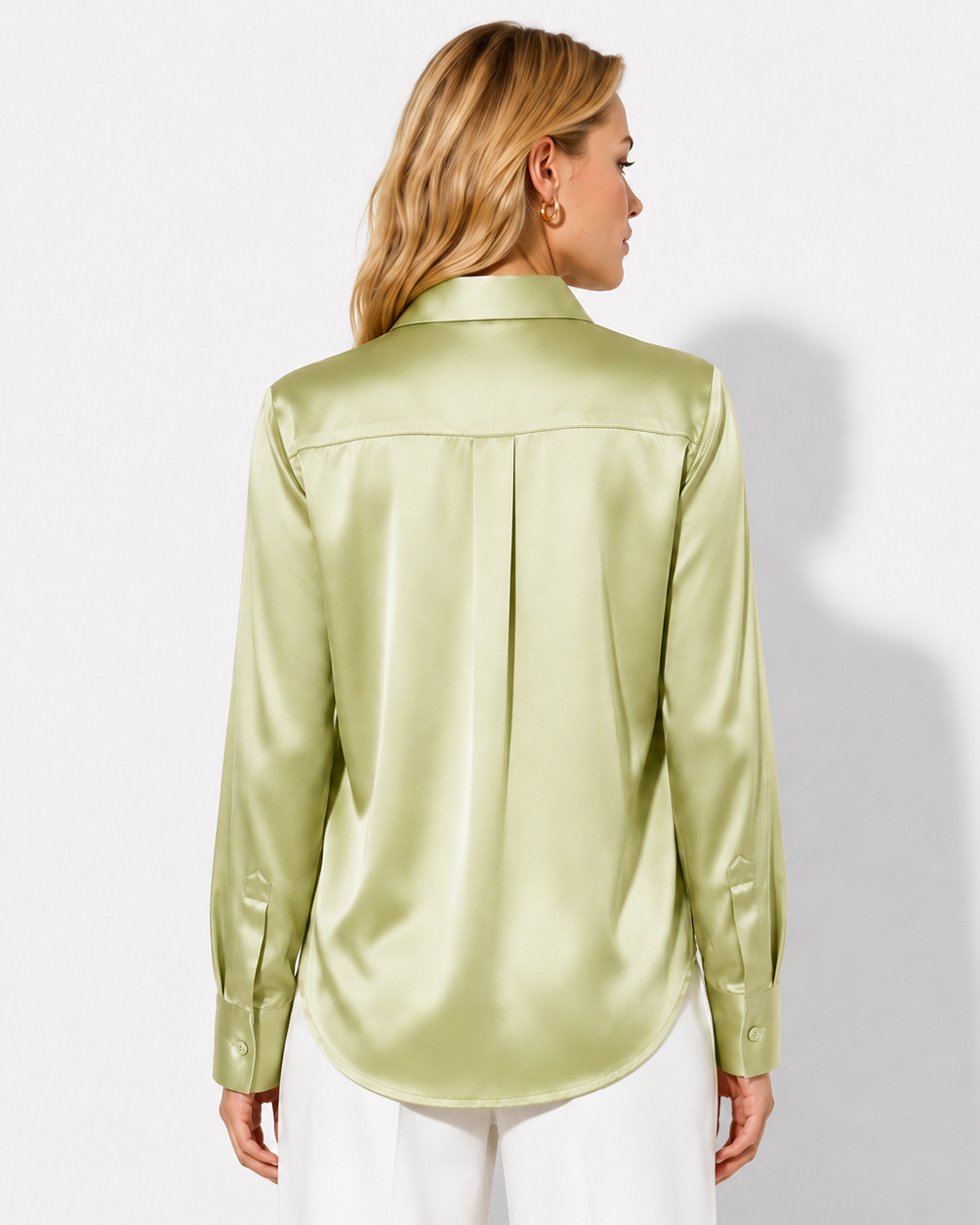A woman shown from the back wearing a light olive green satin button-down shirt and white pants, photographed against a plain light gray studio background.