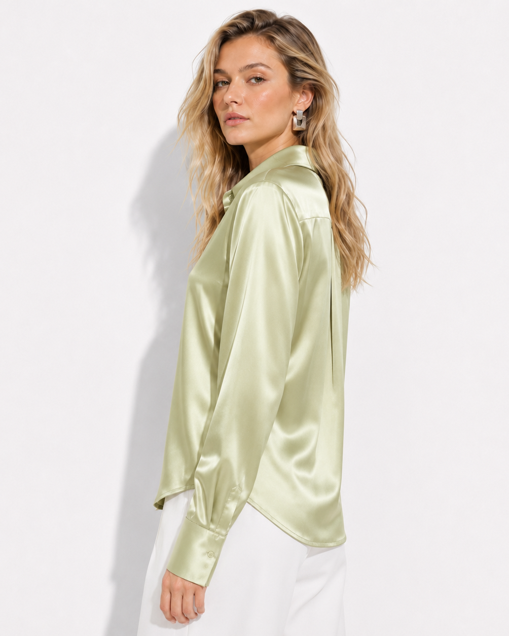 A woman wearing a light sage green satin long-sleeve blouse with a collar, paired with white pants, photographed against a clean white studio background.