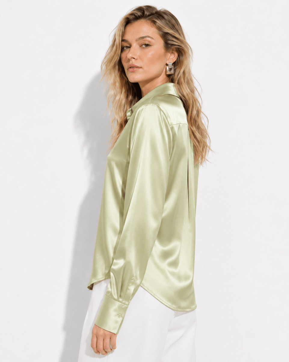 A woman wearing a light sage green satin long-sleeve blouse with a collar, paired with white pants, photographed against a clean white studio background.