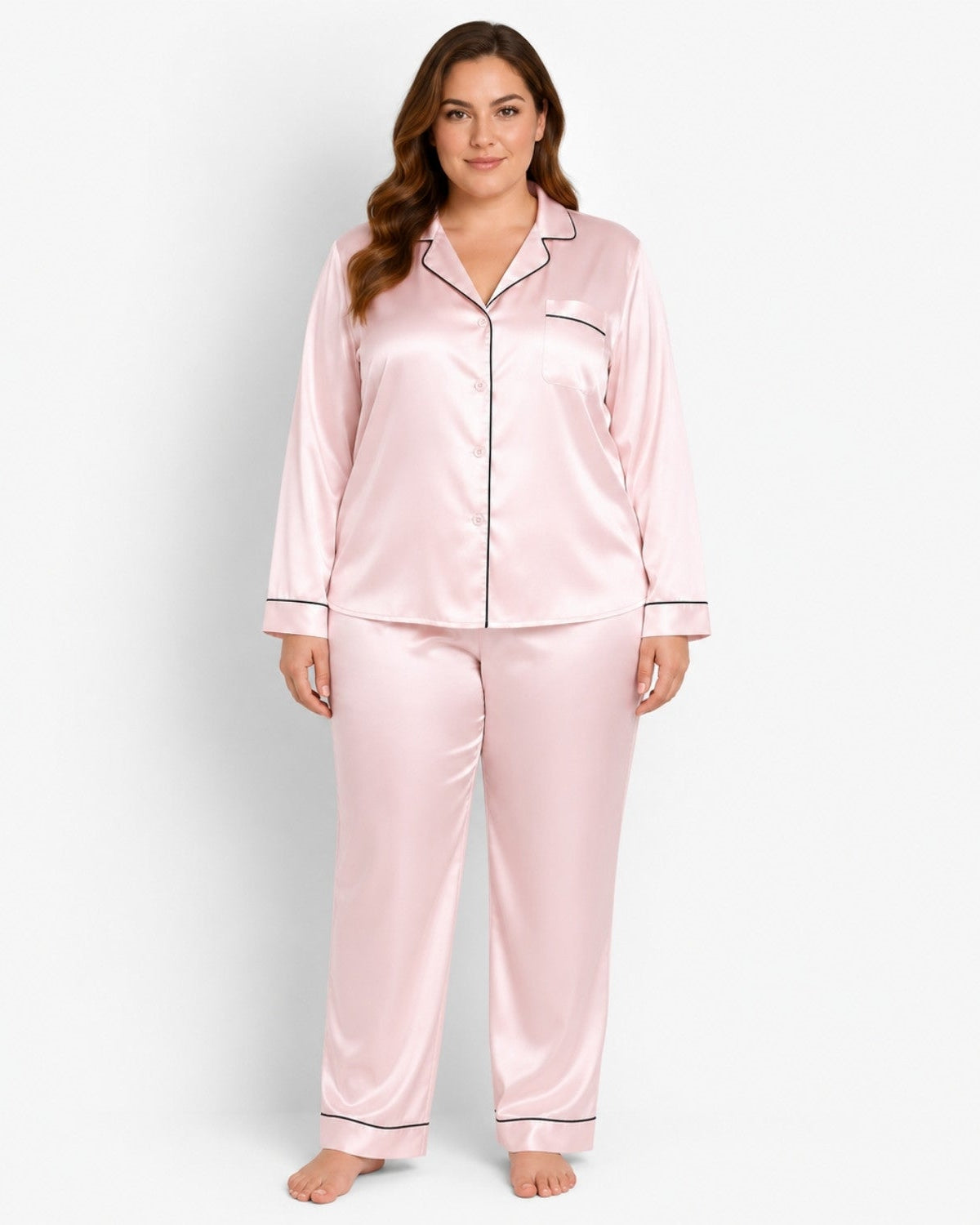 A woman modeling a two-piece satin pajama set in light pink with thin black piping trim, standing against a plain white studio background.