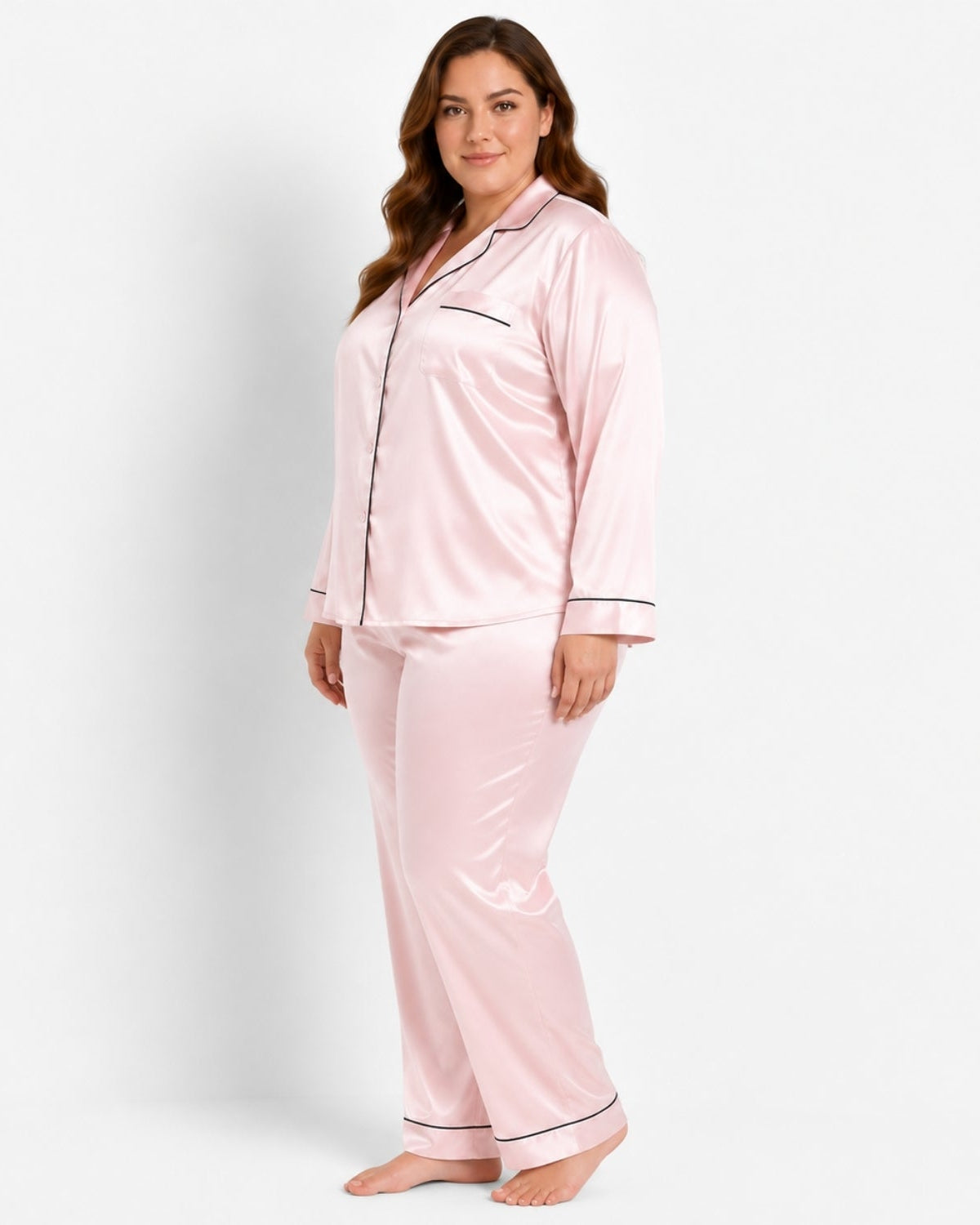 A woman modeling a light pink satin pajama set with black piping details, consisting of a long-sleeve button-up top and matching pants, against a plain white studio background.